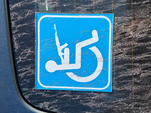 Een groot, liggend stenen beeld van een persoon, half ingebed in een betonnen blok, naast een modern gebouw met geparkeerde auto’s en een heldere lucht.