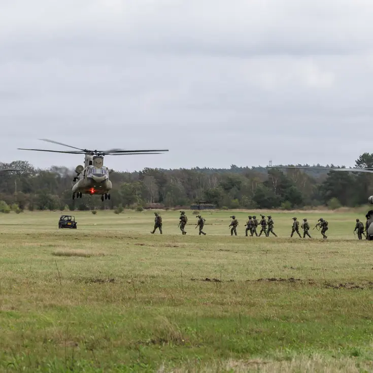 Drie grote militaire helikopters landen in een groen veld, soldaten rennen uit de voorste, voertuigen en bomen op de achtergrond.