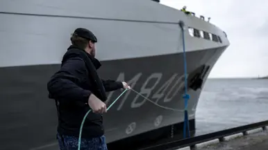 Een militair van de marine haalt met een touw een van de trossen binnen. Daarnaast enkele matrozen en onderofficieren bij de reling op het dek van het schip.