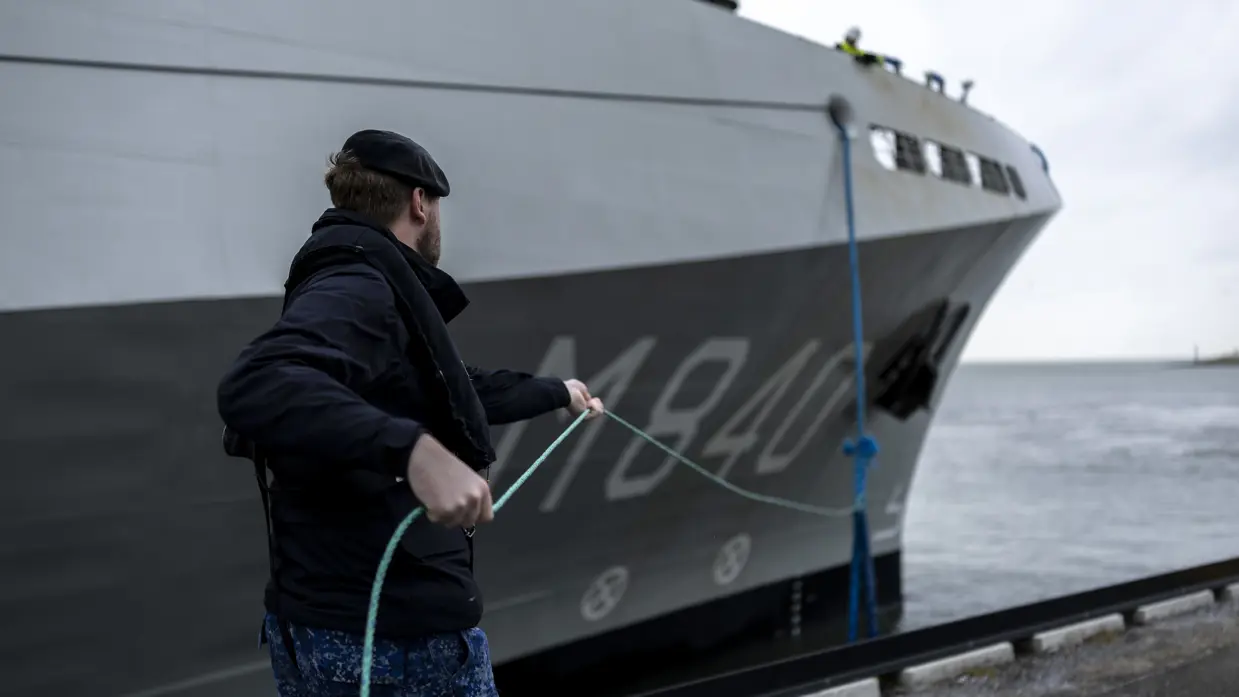 Een militair van de marine haalt met een touw een van de trossen binnen. Daarnaast enkele matrozen en onderofficieren bij de reling op het dek van het schip.
