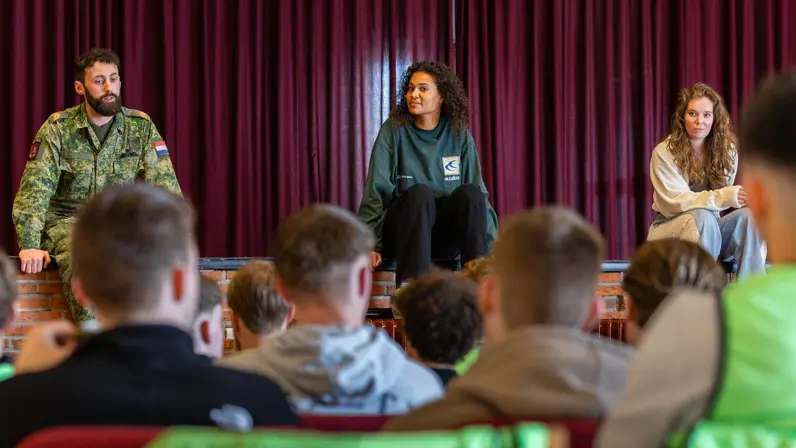 Drie personen zitten op een podium: een militair links, een vrouw in een donkere trui midden, en een vrouw rechts. Een groep jongeren luistert aandachtig in een zaal.