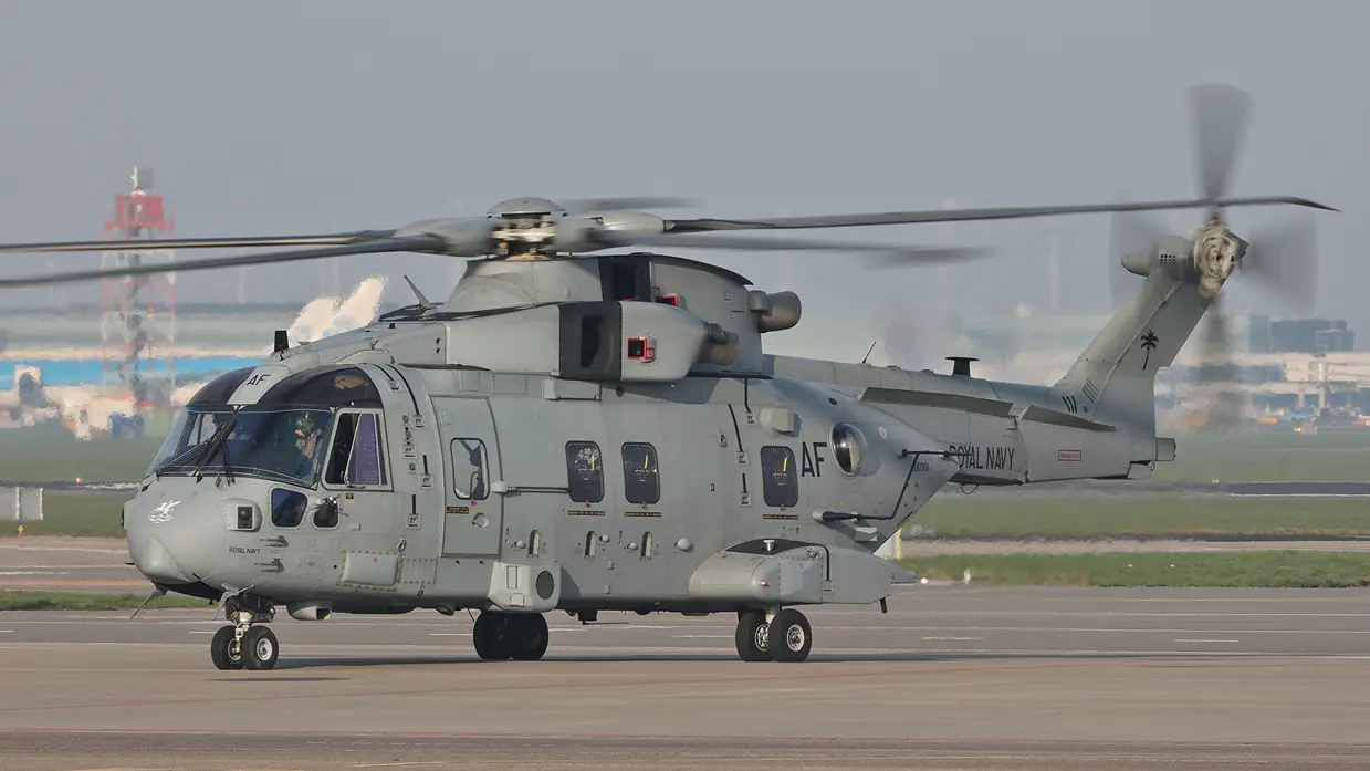  Een grote, grijze militaire helikopter met draaiende rotorbladen staat op een startbaan, met "Amsterdam" en taxiborden op de achtergrond.