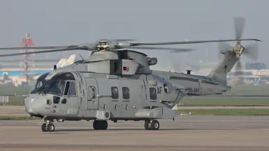  Een grote, grijze militaire helikopter met draaiende rotorbladen staat op een startbaan, met "Amsterdam" en taxiborden op de achtergrond.
