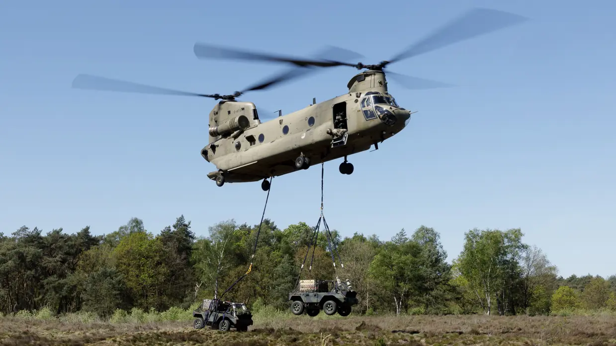 Een militaire helikopter tilt een jeep op boven een bosrijk heideveld, met een andere jeep op de grond. 