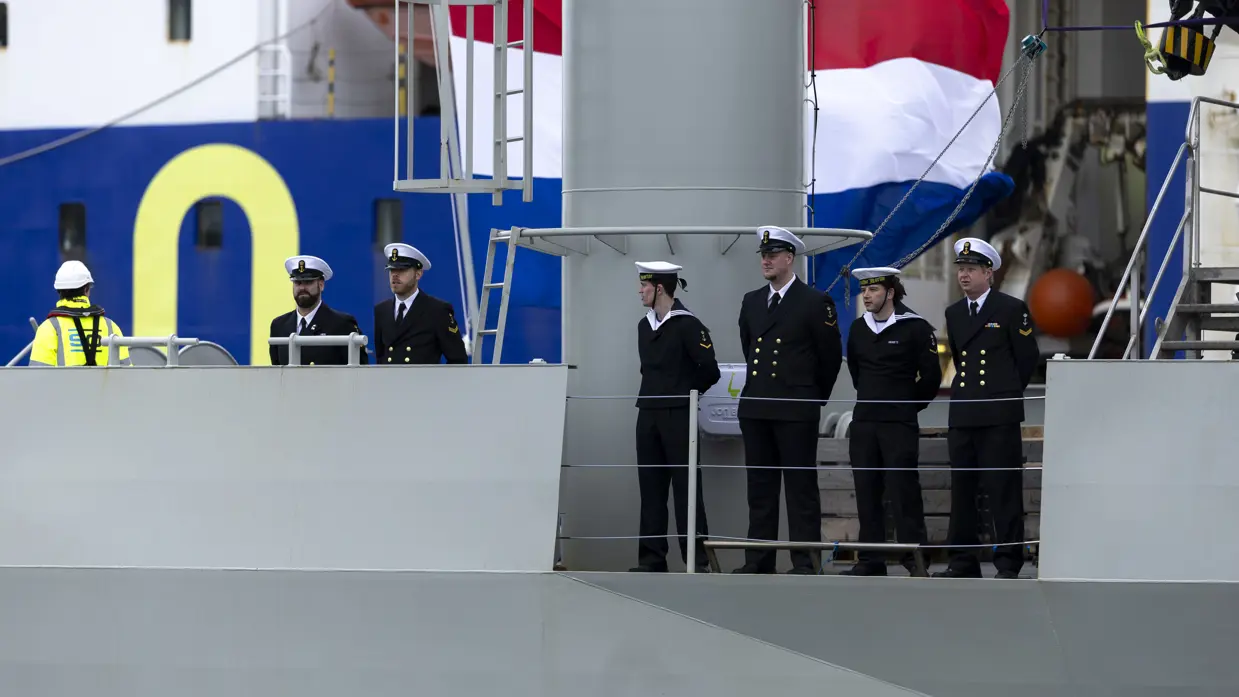 Een militair van de marine haalt met een touw een van de trossen binnen. Daarnaast enkele matrozen en onderofficieren bij de reling op het dek van het schip.