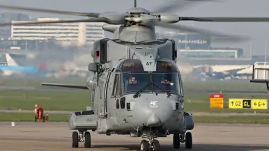  Een grote, grijze militaire helikopter met draaiende rotorbladen staat op een startbaan, met "Amsterdam" en taxiborden op de achtergrond.