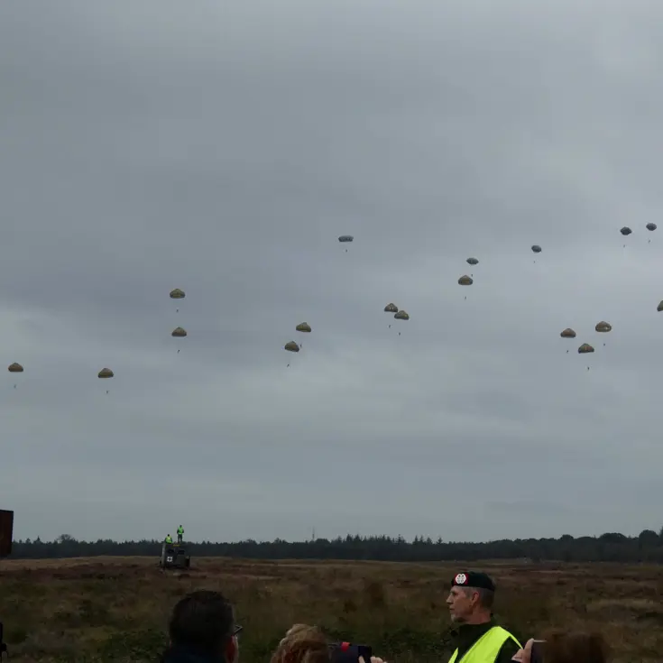  Meerdere para's dalen uit de lucht boven een open veld, toeschouwers kijken toe onder een bewolkte hemel. 