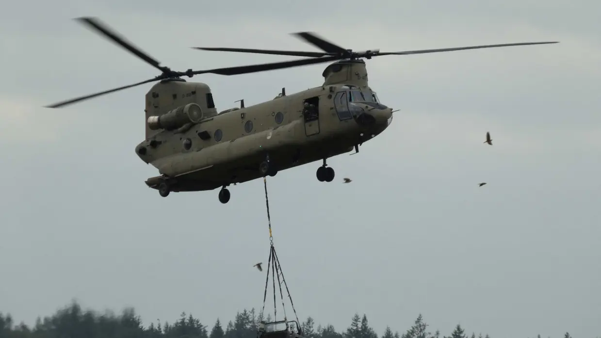 Een militaire helikopter tilt een terreinvoertuig op onder een bewolkte hemel, met bomen en vogels op de achtergrond.