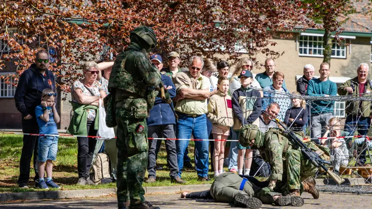 Soldaten in camouflage voeren een oefening uit, omringd door toeschouwers achter prikkeldraad en bomen. 