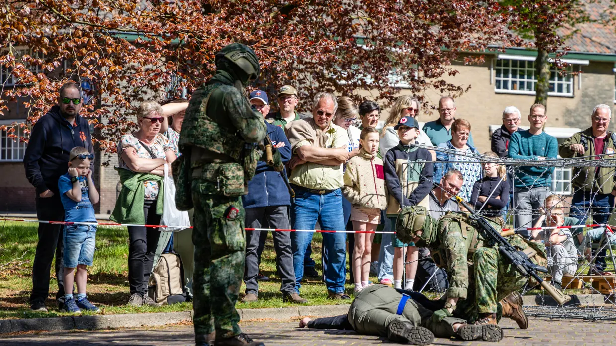 Soldaten in camouflage voeren een oefening uit, omringd door toeschouwers achter prikkeldraad en bomen. 