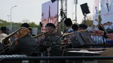 Een buitenoptreden: brassband met trompetten en trombones, microfoons, bladmuziek, publiek op achtergrond, avondlicht en vlaggen. 