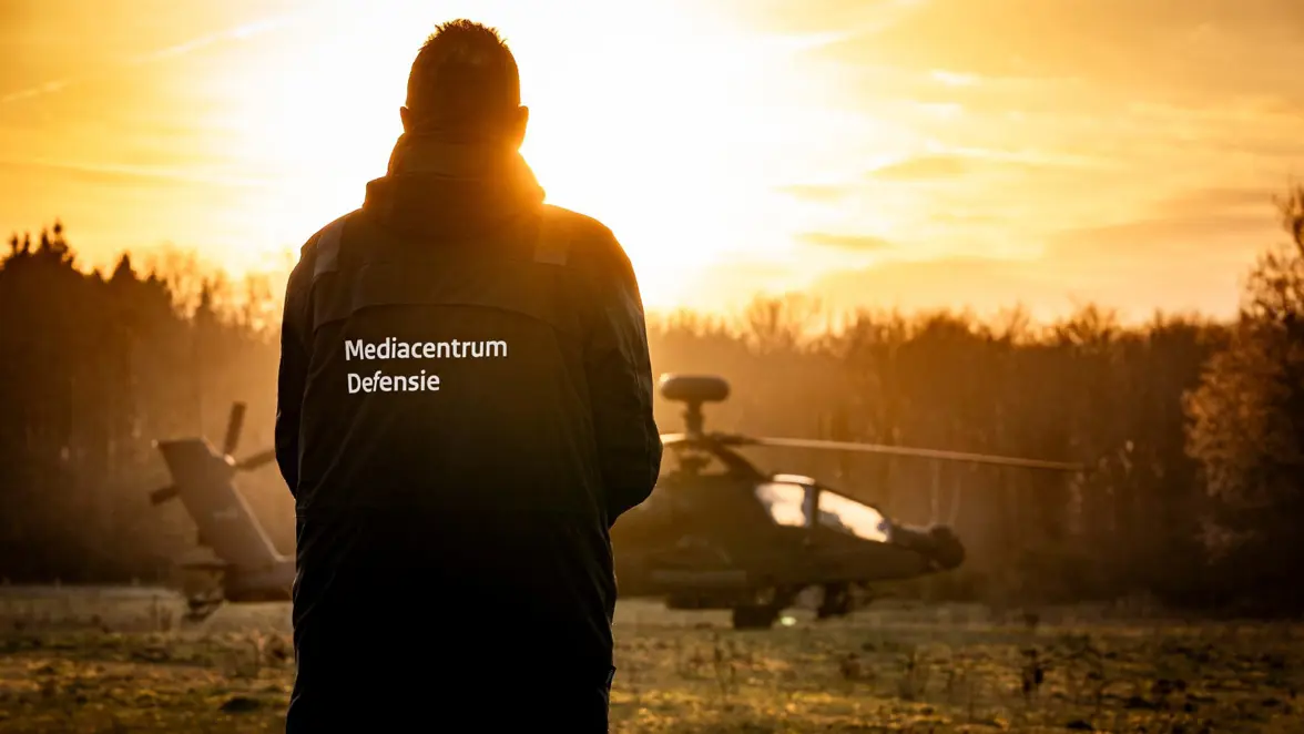 Een persoon van achteren, gekleed in een jas met "Mediacentrum Defensie" erop, staand voor een helikopter tijdens een zonsondergang in een natuurlijke omgeving.