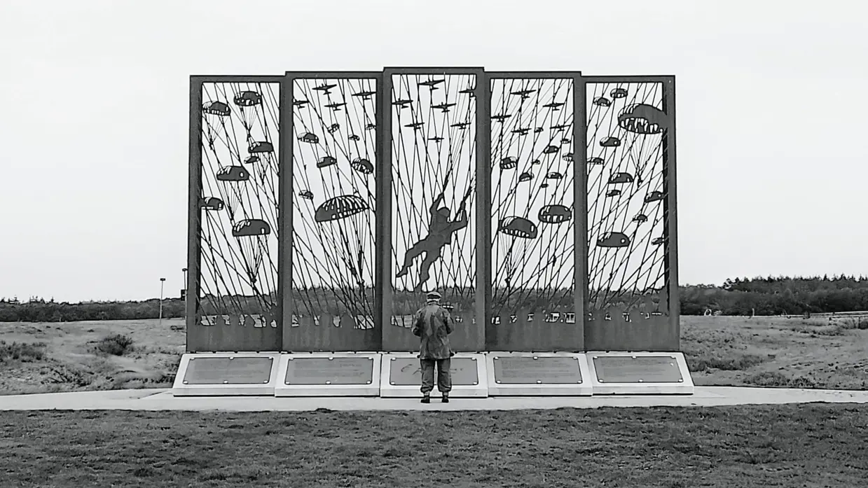 Het silhouet van een man staat voor een groot metalen monument met parachutisten en vliegtuigen, in een open veld.