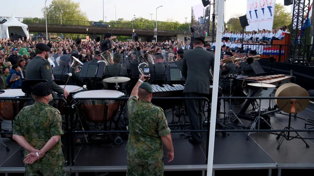 Groot buitenconcert: militaire band speelt op podium, publiek kijkt toe, soldaten in camouflage, drums, trompetten, vlaggen en bomen. 