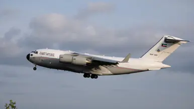 Een wit vliegtuig met de tekst "KUWAIT AIR FORCE" en nummer "KAF 343" vliegt door een bewolkte lucht. 