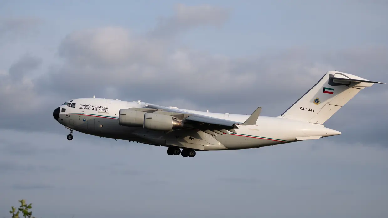 Een wit vliegtuig met de tekst "KUWAIT AIR FORCE" en nummer "KAF 343" vliegt door een bewolkte lucht. 