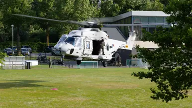 Een witte militaire helikopter met open deur, twee bemanningsleden, staat op een grasveld bij een gebouw en bomen. 