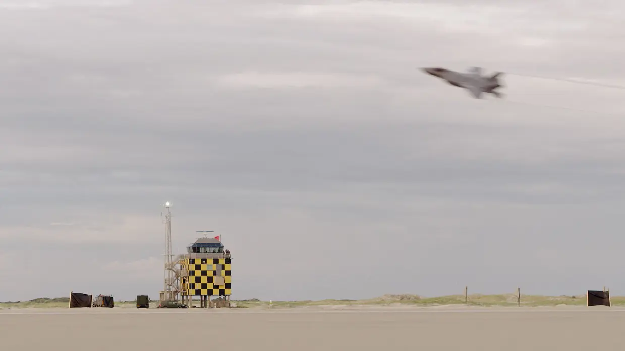 Een F-35 vliegt schuin over strand en laag duinlandschap. Op het strand staat een zwart-geel geblokte gevechtsleidingstoren op palen. Ernaast een zendmast en een paar groene legervoertuigen.