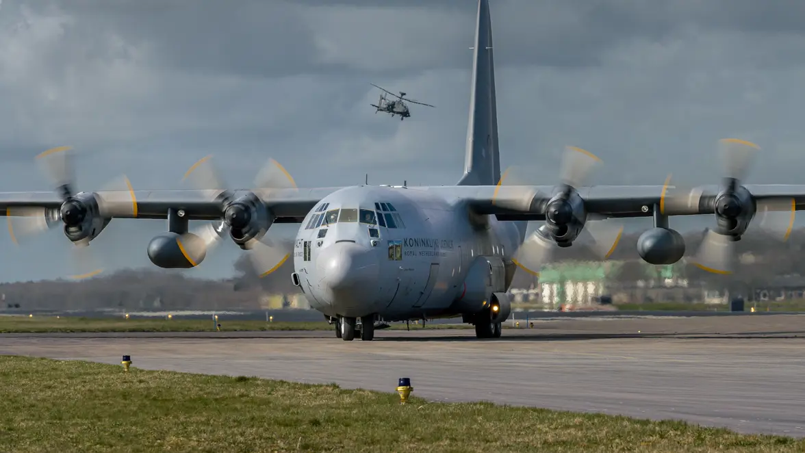 Een groot militair transportvliegtuig met vier draaiende propellers op een startbaan, terwijl een helikopter er boven vliegt tegen een bewolkte lucht.
