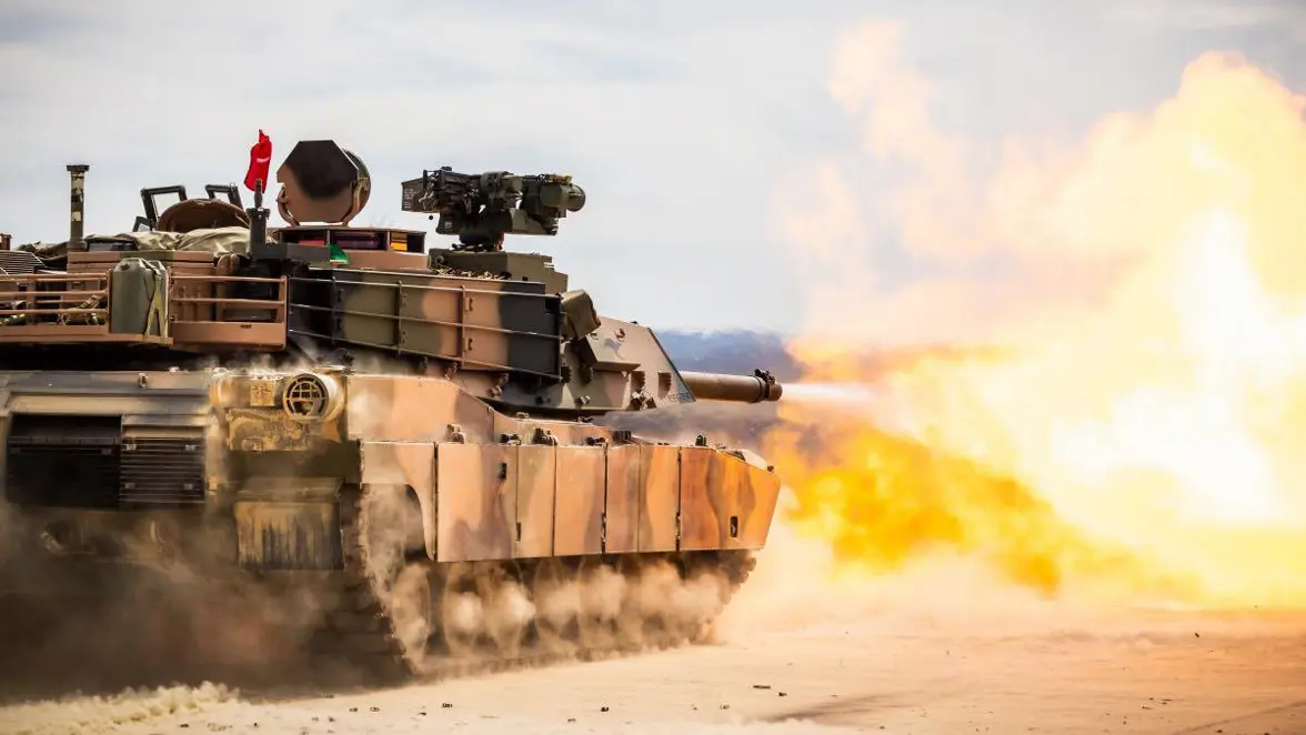 Een tank vuurt zijn kanon af in een woestijn, met zand en vuur dat uit de loop spat, terwijl de tank in beweging is.