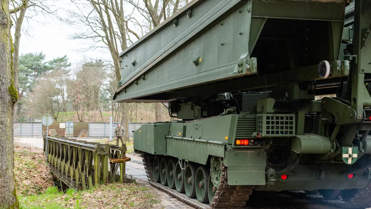 +alttekst: Een groene bruglegger legervoertuig met rupsbanden plaatst een zware metalen brug over een pad, omringd door bomen en een hek. 