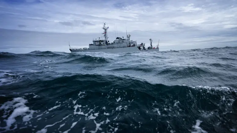 Een groot, grijs schip vaart door woeste, donkerblauwe golven onder een bewolkte, grijze hemel, met witte schuimkoppen op het water.