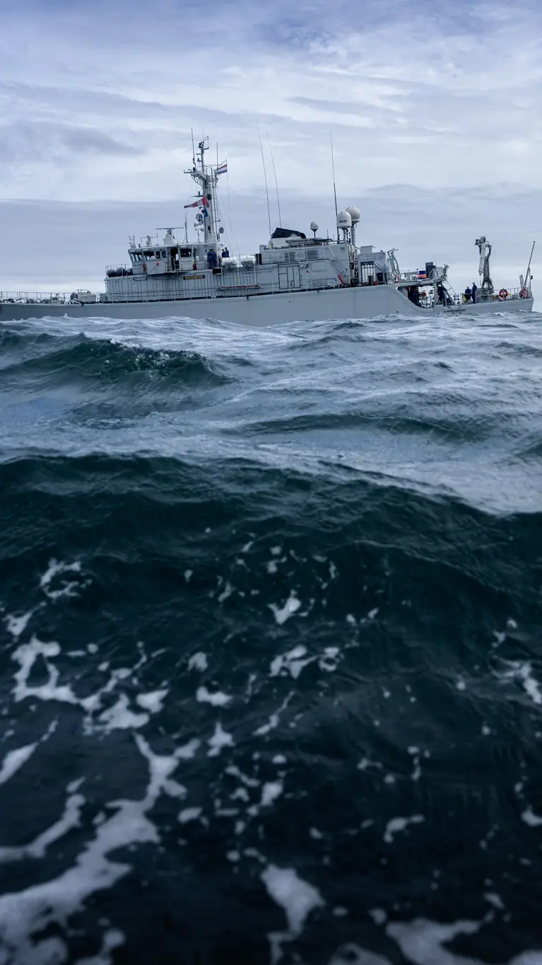 Een groot, grijs schip vaart door woeste, donkerblauwe golven onder een bewolkte, grijze hemel, met witte schuimkoppen op het water.