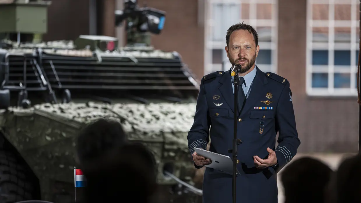 Een militair van de luchtmacht spreekt een zaal toe met achter hem een militair voertuig.