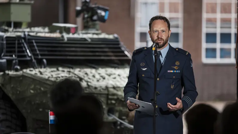 Een militair van de luchtmacht spreekt een zaal toe met achter hem een militair voertuig.