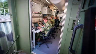 Vier personen, van wie twee in militair uniform, zitten achter beeldschermen in een groene container.