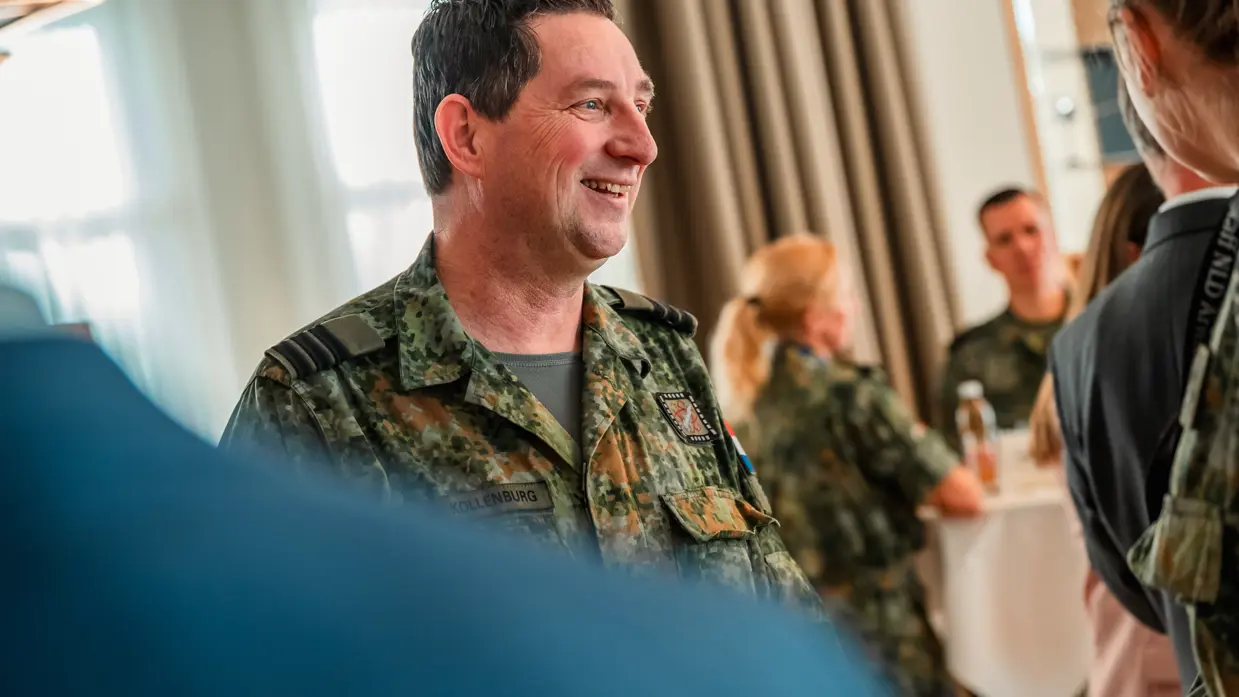 Een lachende militair met op de achtergrond 2 collega’s aan een tafel. 