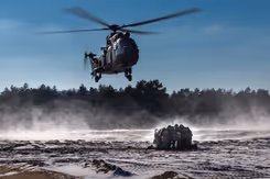 Legerhelikopter hangt net boven de grond terwijl groepje militairen ineengedoken bij elkaar zit om zich te beschermen tegen wind en stof.