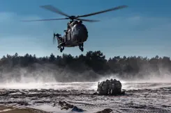 Legerhelikopter hangt net boven de grond terwijl groepje militairen ineengedoken bij elkaar zit om zich te beschermen tegen wind en stof.