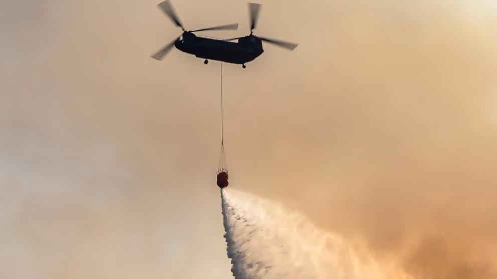 Chinook-transporthelikopter in oranjegekleurde lucht stort water uit met bambi-bucket onder het toestel.