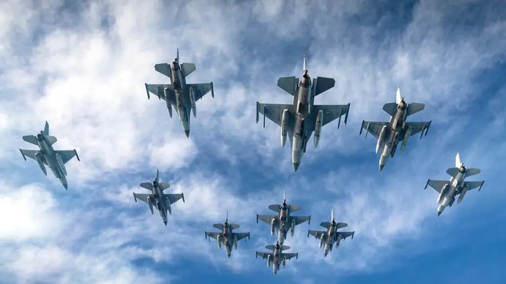 Een formatie F-16 vliegtuigen van onderaf gezien. Een blauwe lucht op de achtergrond met enkele wolken.