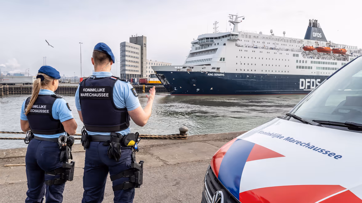 Marechausseevoertuig met 2 marechaussees ervoor. Zij kijken naar een schip in de haven.