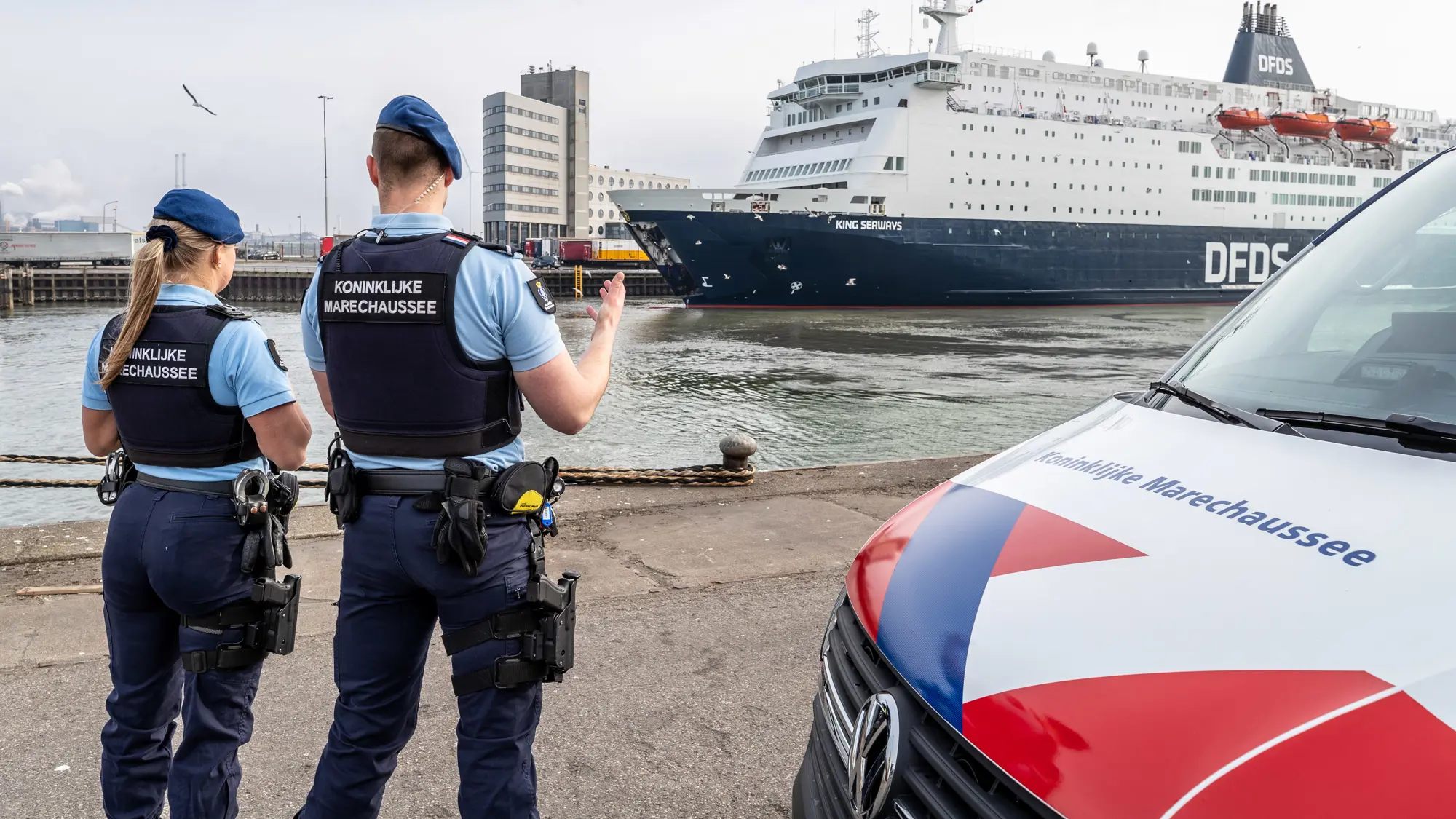 Marechausseevoertuig met 2 marechaussees ervoor. Zij kijken naar een schip in de haven.