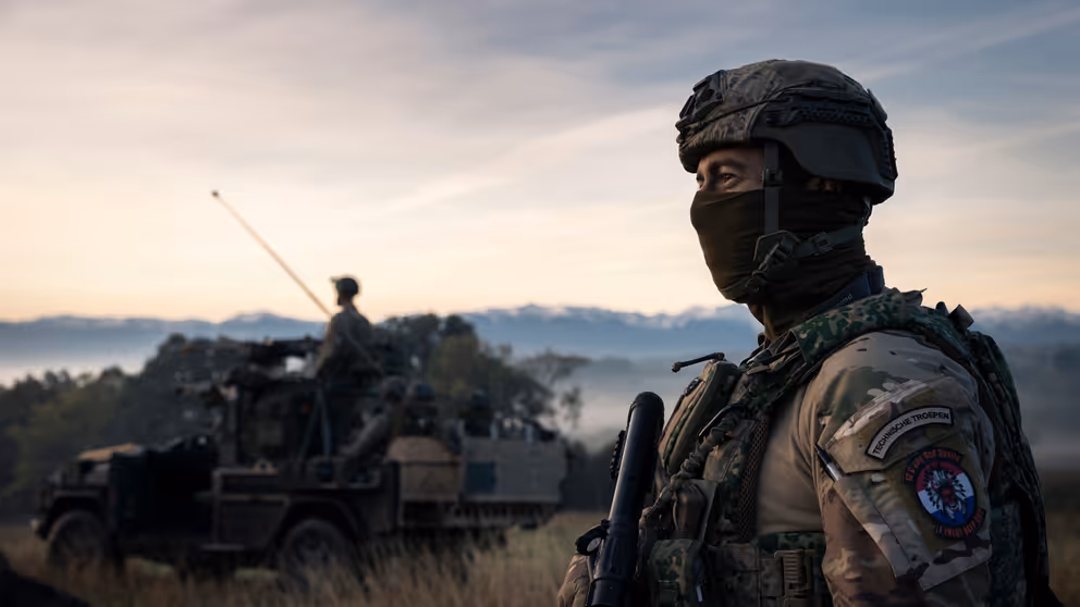 Militair met gezichtsbedekking op de uitkijk naast militair voertuig in natuurgebied.
