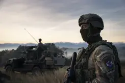 Militair met gezichtsbedekking op de uitkijk naast militair voertuig in natuurgebied.
