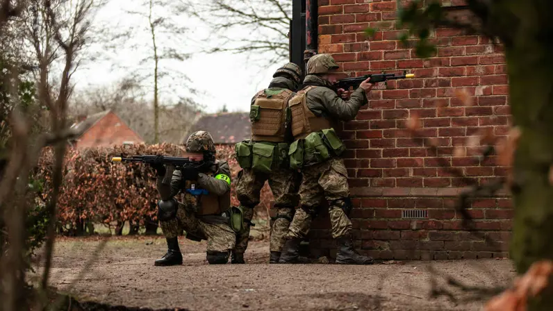3 soldaten in camouflage richten hun wapens. Ze staan op hoek van gebouw met rode bakstenen, omringd door kale bomen en struiken.