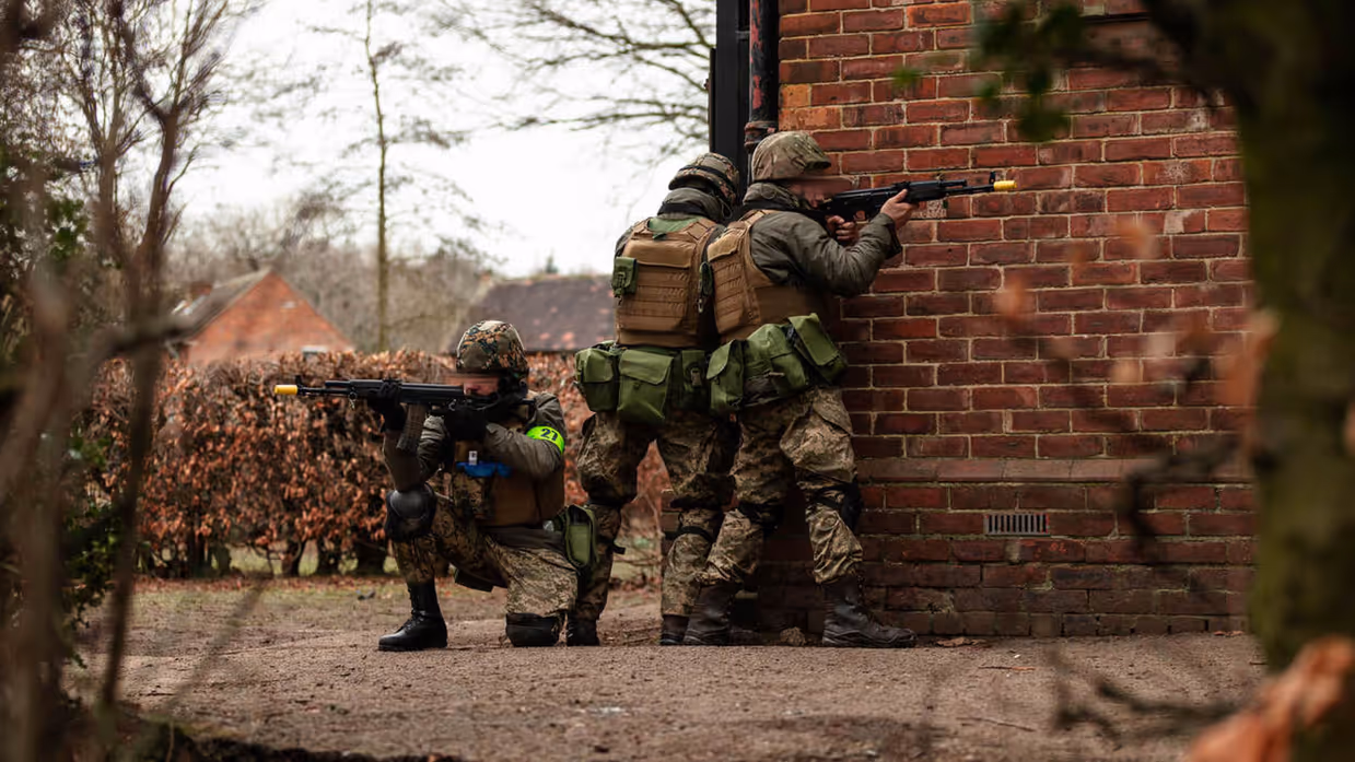3 soldaten in camouflage richten hun wapens. Ze staan op hoek van gebouw met rode bakstenen, omringd door kale bomen en struiken.