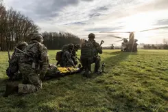 5 militairen zitten op hun knieën in het gras bij een gewonde op een brancard. 2 Chinook-transporthelikopters zijn zichtbaar op de achtergrond.