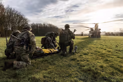 Militairen oefenen op een grasveld. 1 ligt op een brancard. Op de achtergrond 2 Chinook-transporthelikopters.
