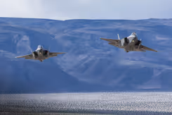 2 F-35 jachtvliegtuigen vliegen boven water tegen een achtergrond van bergen in blauw licht.