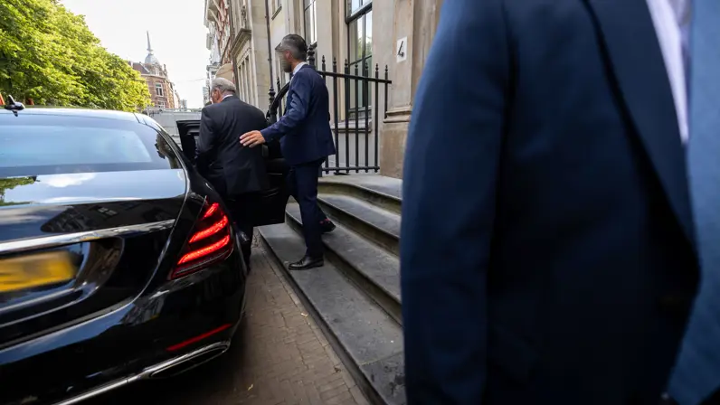 en man in donkerblauw pak begeleidt een oudere man een auto in. Dit voertuig heeft geblindeerde ramen.