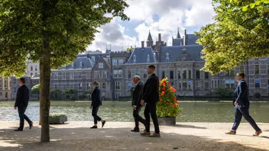 Drie persoonsbeveiligers staan in een gang en omringen een bellende man in pak. Daarnaast vier persoonsbeveiligers die rondom een man lopen bij het Binnenhof in Den Haag.