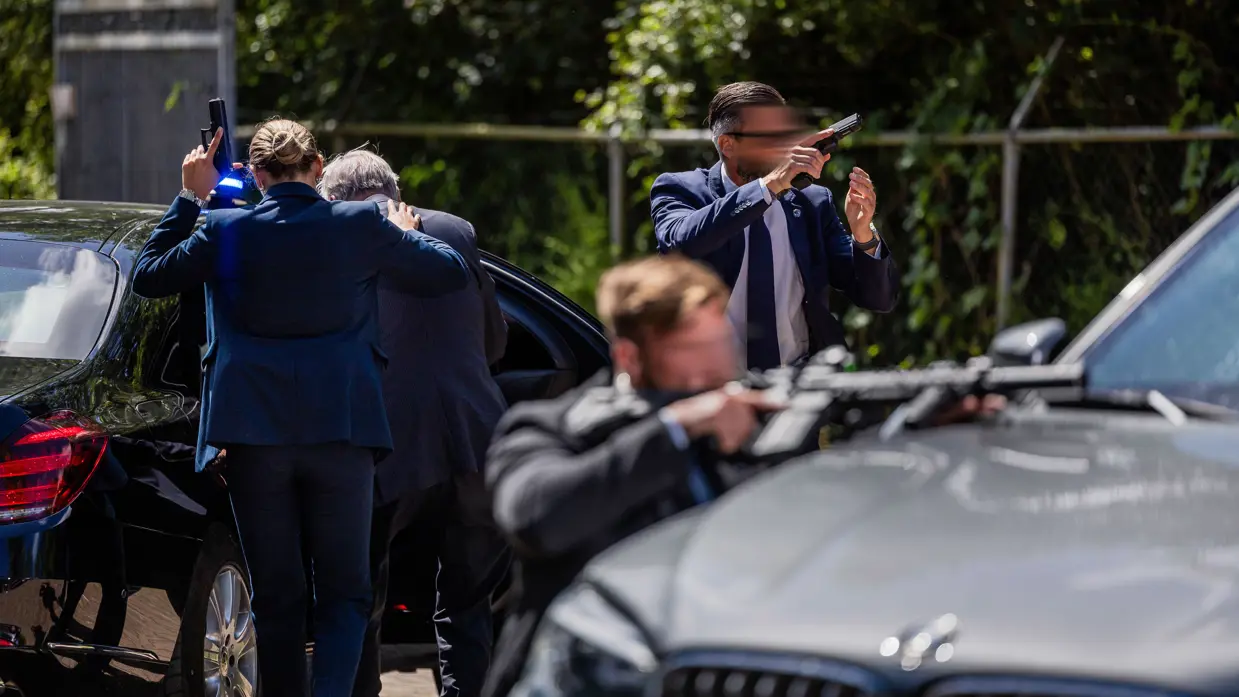 Drie persoonsbeveiligers achter een auto. Een van hen begeleidt de TBP de auto in, haar pistool in de lucht gericht. De twee daarvoor richten de andere kant op. De een met een pistool, de ander met een lang wapen.