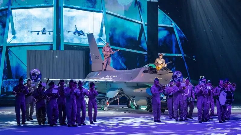 Op een blauw-paars verlicht podium bespelen muzikanten van de luchtmacht op en rondom een F-16 hun instrument.