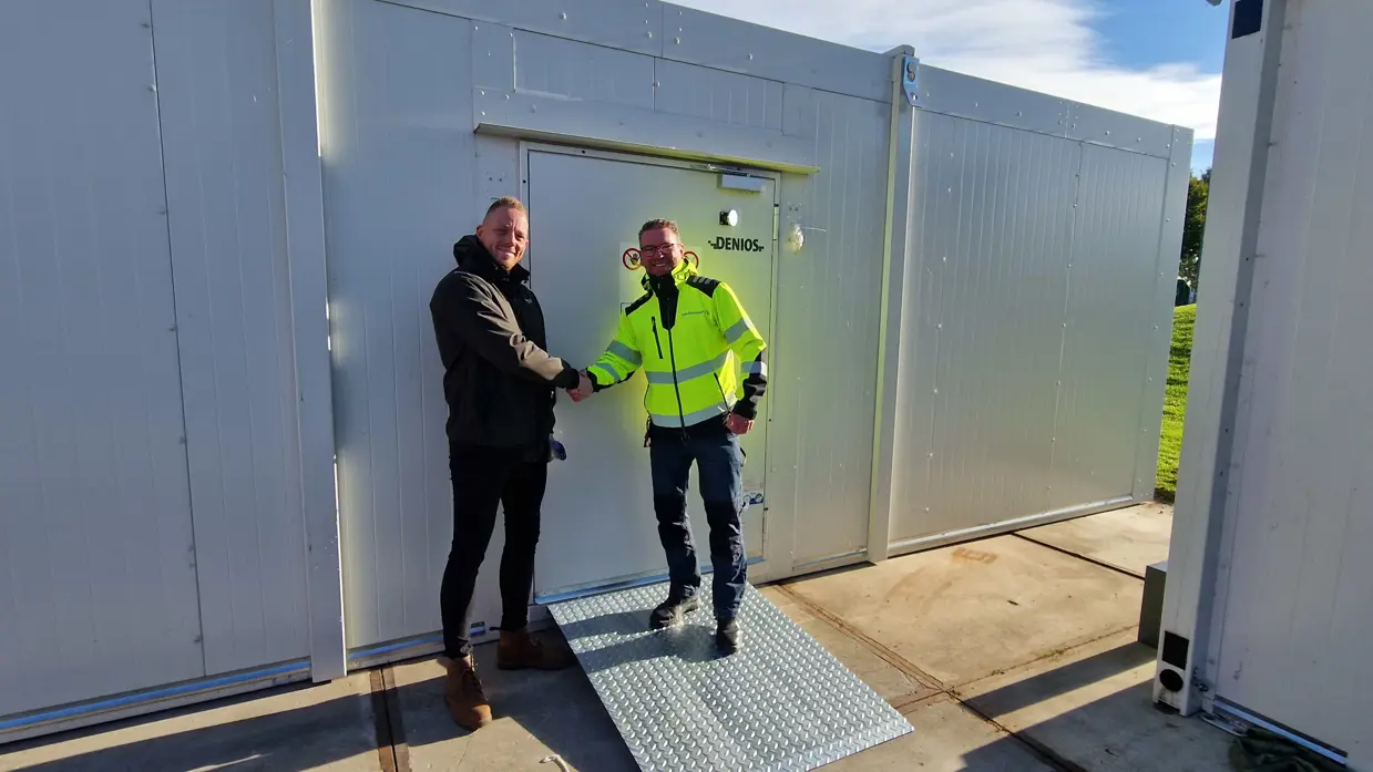Glenn Afman in zwarte winterjas schudt de hand van een man in felgele werkjas. Ze staan buiten voor en tussen witte containers.
