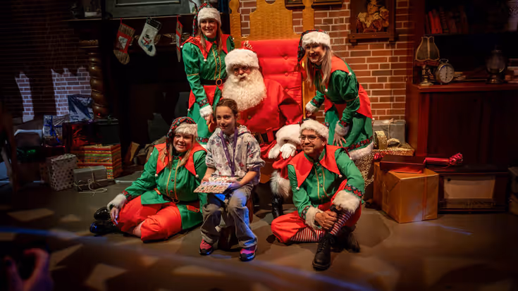 De kerstman poseert met een kindje voor een fotograaf, omgeven door helpers van de kerstman.